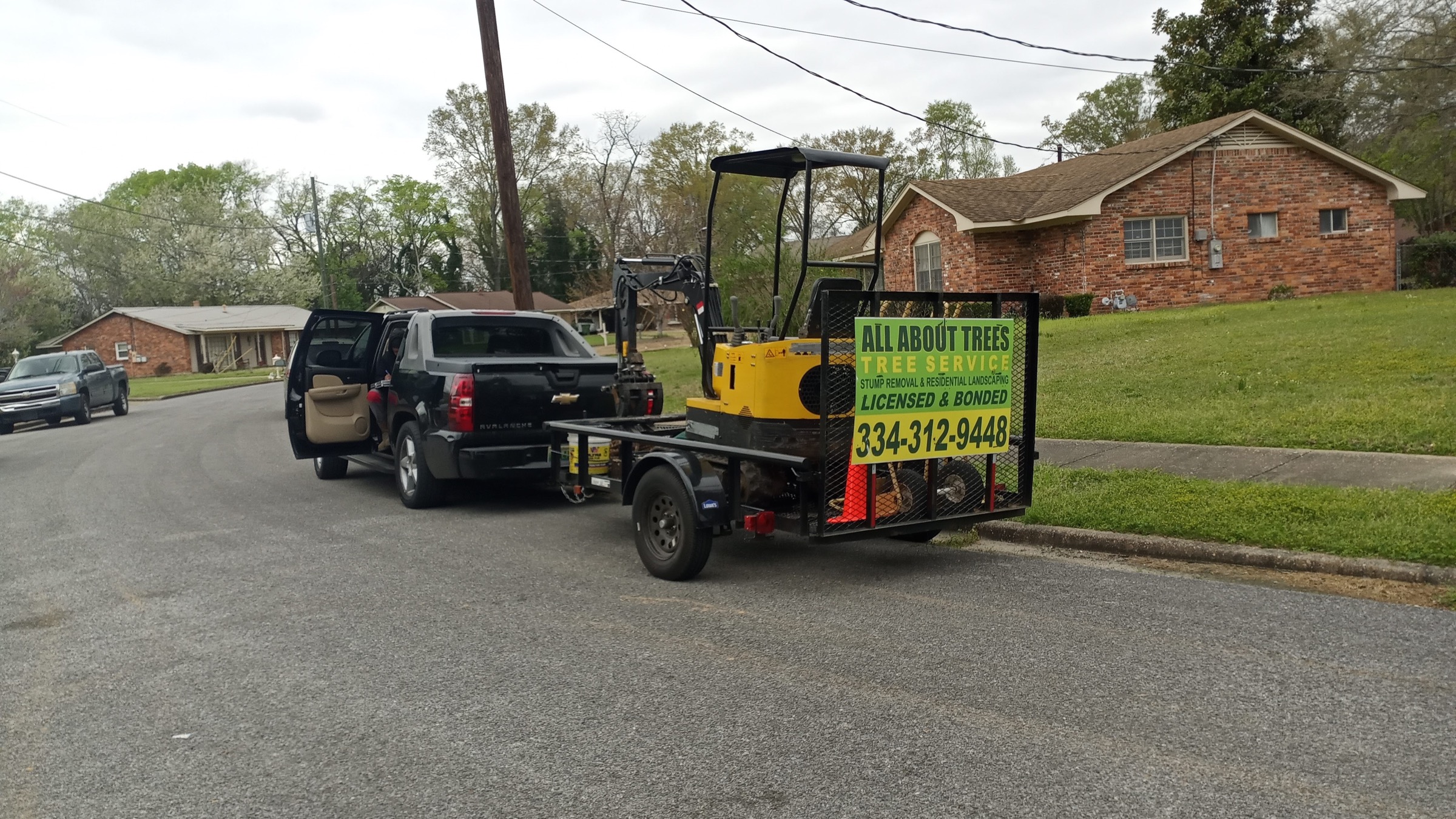 The All About Trees truck and trailer on a job, loaded with the tracked machine, the sign reading Licensed & Bonded